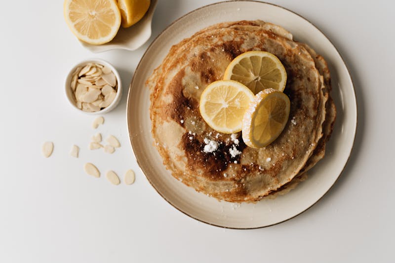 A mouth-watering stack of pancakes topped with lemon slices and surrounded by almond flakes.