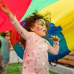 Children enjoying a lively outdoor party under vibrant parachute play, face painted with colorful designs.